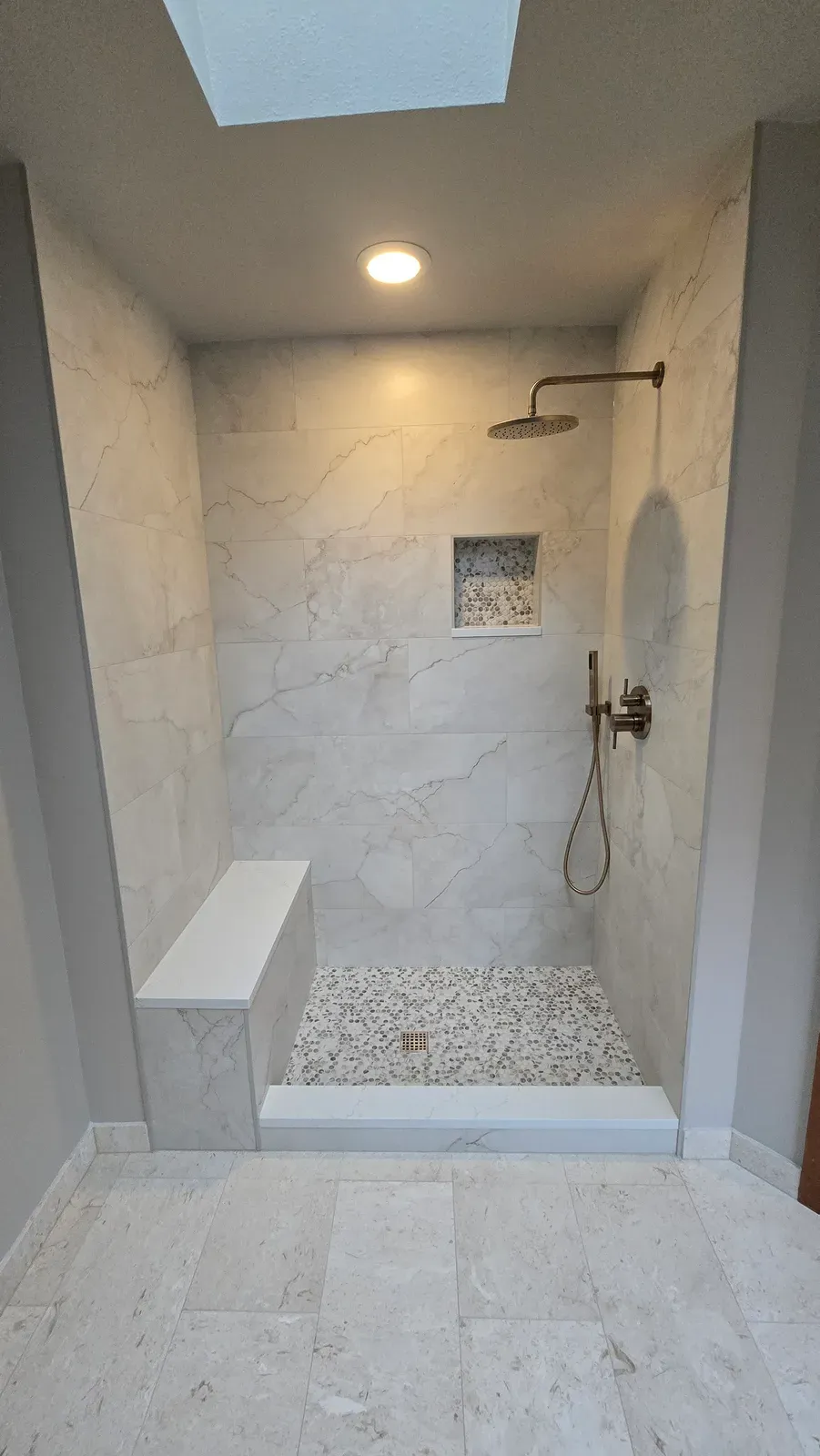 walk-in shower with a bench, pebble floor, rain head and handheld under a skylight